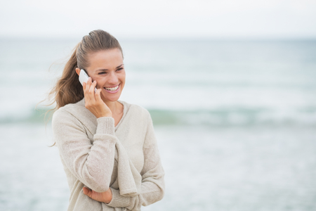 Smiling young woman talking cell phone on cold beachの写真素材