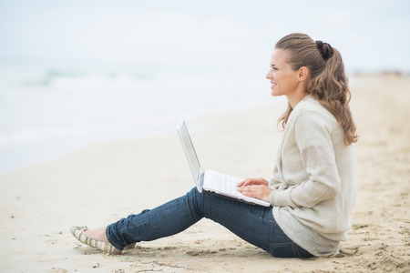 Happy young woman sitting with laptop on cold beachの写真素材