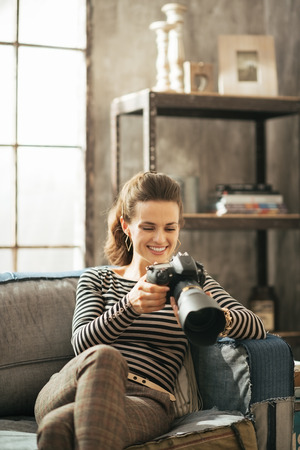 Happy young woman sitting on divan and using modern dslr photo cameraの写真素材
