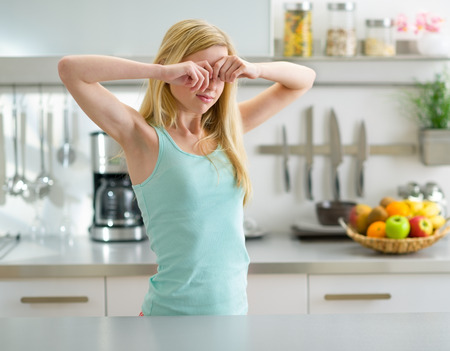 Young woman standing in in kitchen and rubbing eyes after sleepの写真素材