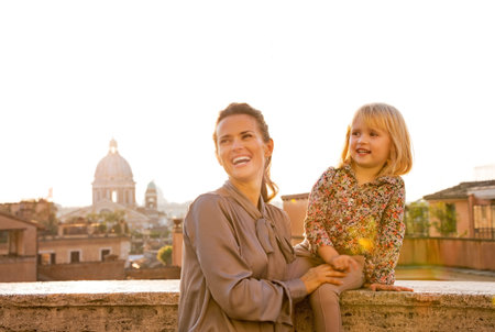 Mother and baby girl on street overlooking rooftops of rome on sunset looking into distanceの写真素材
