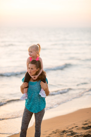 Happy baby girl sitting on shoulders of mother on beach in the evening and looking into distanceの写真素材