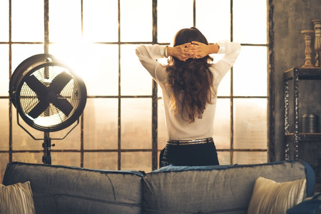 Elegant brunette woman seen from behind, leaning against the back of a sofa in a loft. Hands laced behind her head, she is looking out the loft window. Industrial chic atmosphere.の写真素材