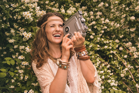 Longhaired hippy-looking young lady in knitted shawl and white blouse standing among flowers playing with retro cameraの写真素材
