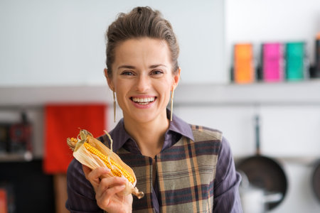 A happy, smiling woman is holding a corn on the cob with its husk. She's excited about the cornbread...