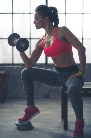 A fit, muscular woman sitting on a city loft gym bench is lifting weights, resting her elbow against her knee, curling the weights towards her as she looks down at her hand.の写真素材