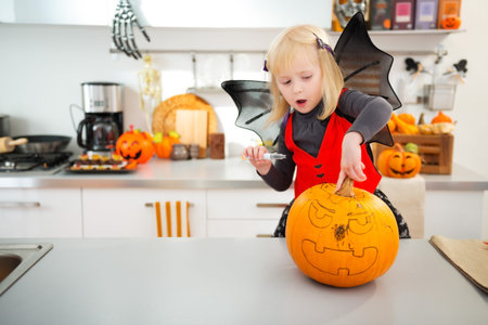 Interested blond girl in bat costume using stencils to carve big orange pumpkin Jack-O-Lantern on Halloween party in decorated kitchen. Traditional autumn holidayの写真素材