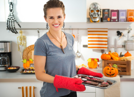 Smiling modern housewife in decorated kitchen holding tray with freshly baked Halloween biscuits for Trick or Treat. Ready to halloween party. Traditional autumn holidayの写真素材