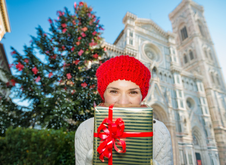 Cheerful woman tourist in winter outfit hiding behind present gift box while standing near large Christmas tree in Florence's historic center. Christmas and travel concept.の写真素材