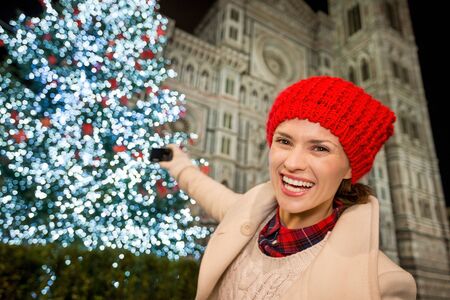 Happy young woman in white coat taking photo of Christmas tree near Duomo in the evening. She having holiday tour and enjoying Christmas decorated historical area of Florence, Italyの写真素材