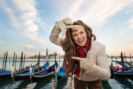 Sunset brings to life irresistible magic of Venice - the unique Italian city. Happy young woman traveler standing on embankment in the front of the line of gondolas and framing with handsの写真素材