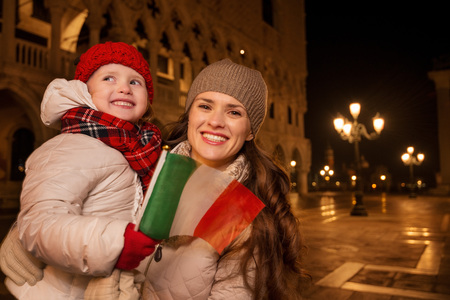 Holiday family trip to Venice, Italy can change the whole Christmas experience. Happy mother and child holding Italian flag standing on Piazza San Marco in the evening. Winter Tourismの写真素材