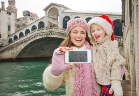 Small family trip is another way how to make Christmas holidays even more appealing. Happy mother and child in Santa Hat taking selfie with smartphone in front of Ponte di Rialtoの写真素材
