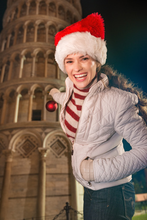 Portrait of smiling young woman in Santa hat decorating Leaning Tower of Pisa with Christmas ball. Italy.の写真素材