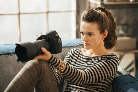 Young brunet woman is sitting on couch and holding dslr photo camera