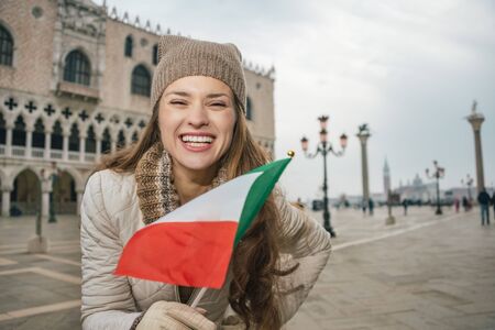 Delightful Venice, Italy can help make the most of your next winter getaway. Portrait of happy young woman tourist with Italian flag standing on St. Mark's Square near Dogi Palaceの写真素材