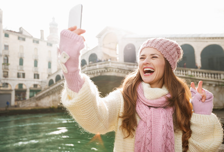 Ultimate getaway shortcut - start New Year going on Carnival in Venice, Italy. Happy woman tourist in white sweater showing victory while taking selfie with smartphone in the front of Rialto Bridgeの写真素材