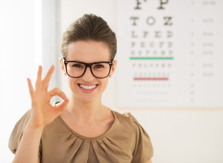 Modern health care. Happy young woman wearing eyeglasses showing ok gesture in front of Snellen...