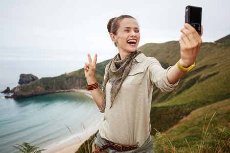 Into the wild in Spain. healthy woman hiker taking selfie with digital camera and showing victory in front of ocean view landscapeの写真素材