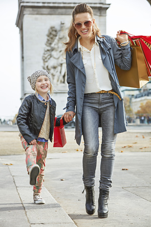 Stylish autumn in Paris. Full length portrait of smiling young mother and child with shopping bags near Arc de Triomphe in Paris, France going forwardの写真素材