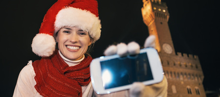 Trip full of inspiration at Christmas time in Florence. Portrait of happy young woman in Christmas hat against Palazzo Vecchio in Florence, Italy showing smartphone's blank screenの写真素材