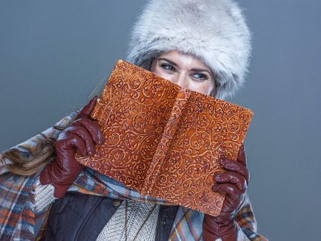 Winter things. Portrait of trendy woman in fur hat isolated on cold blue background hiding behind book and looking copy spaceの写真素材