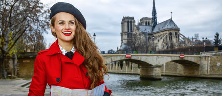 Bright in Paris. smiling elegant tourist woman in red trench coat on embankment near Notre Dame de Paris in Paris, France with map looking into the distanceの写真素材