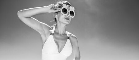 Heading to white sand blue sea paradise. Woman in white swimsuit and funky pineapple glasses looking into the distance at sandy beach on a sunny dayの写真素材