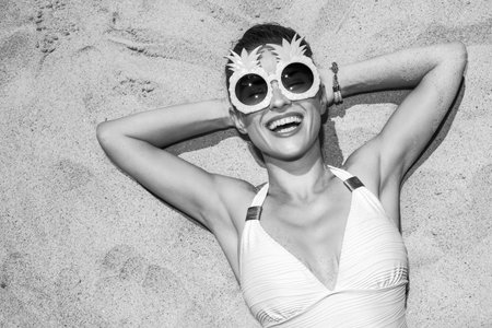 Warm sand treatment. smiling woman in swimsuit and funny pineapple glasses laying on the sand
