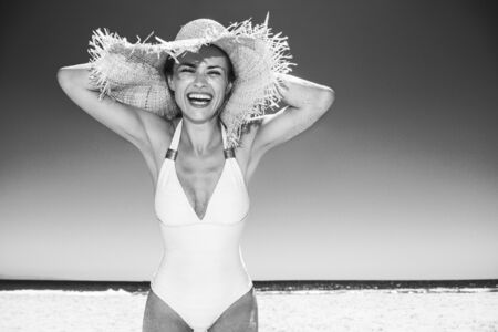 Heading to white sand blue sea paradise. Portrait of smiling woman in white swimsuit and straw hat at sandy beach on a sunny dayの写真素材