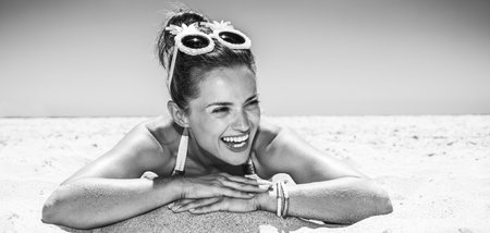 Heading to white sand blue sea paradise. Smiling woman in white swimsuit and funky pineapple glasses laying on sandy beach on a sunny day and looking into the distanceの写真素材