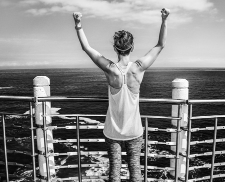 Look Good, Feel great! Seen from behind young healthy woman in fitness outfit rejoicing at the embankmentの写真素材