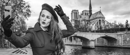 Bright in Paris. cheerful trendy traveller woman in red trench coat on embankment near Notre Dame de Paris in Paris, France with mobile phone taking selfieの写真素材