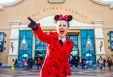 DISNEYLAND, FRANCE - DECEMBER, 8, 2016:. happy modern woman in red trench coat in the front of Disney Studio 1 pointing at somethingのeditorial素材