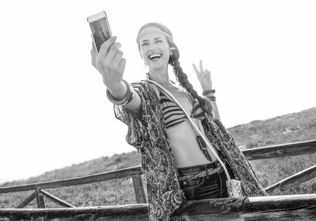 Bohemian vibe vacation. smiling modern free spirit girl in jeans shorts and cape outdoors in the summer evening with smartphone taking selfie and showing victory gestureの写真素材
