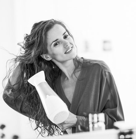 Happy young woman blow drying hair in bathroomの写真素材