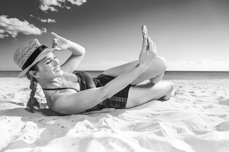 Sun kissed beauty. happy active woman in swimwear on the seacoast with mobile phone taking selfieの写真素材