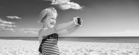 Sun kissed beauty. happy blond child in swimwear on the seacoast with digital camera taking photoの写真素材