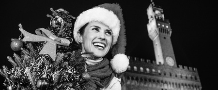 Trip full of inspiration at Christmas time in Florence. Portrait of smiling modern woman in Christmas hat in the front of Palazzo Vecchio in Florence, Italy with Christmas tree looking into the distanceの写真素材