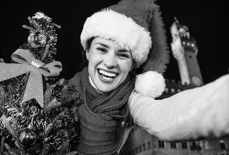 Trip full of inspiration at Christmas time in Florence. happy modern tourist woman in Christmas hat in the front of Palazzo Vecchio in Florence, Italy with Christmas tree taking selfieの写真素材
