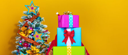 Festive season. stylish woman in colorful clothes near Christmas tree isolated on yellow background holding pile of Christmas present boxesの写真素材
