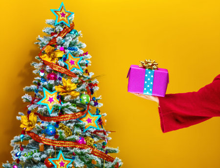 Festive season. Closeup on young woman in colorful clothes near Christmas tree isolated on yellow background showing Christmas present boxの写真素材