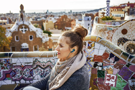 Barcelona signature style. modern traveller woman in coat at Guell Park in Barcelona, Spain listening audioguide while sitting on benchの写真素材