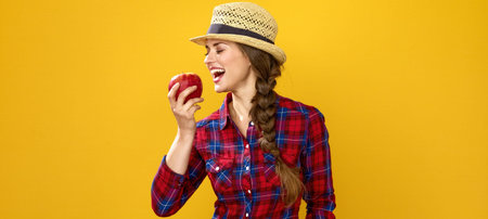 Healthy food to your table. Portrait of smiling young woman grower in checkered shirt on yellow background eating an appleの写真素材