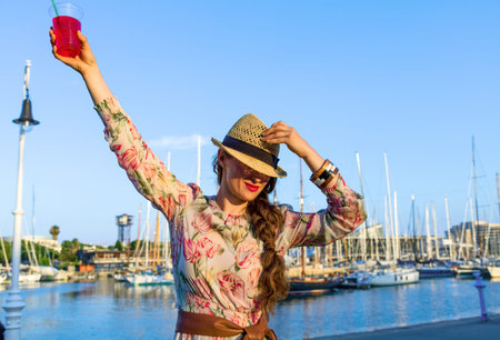 Summertime at colorful Barcelona. stylish traveller woman in long dress and straw hat in Barcelona, Spain with bright red beverage having fun timeの写真素材