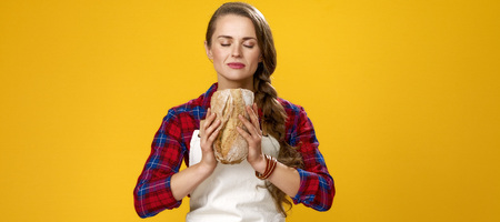 Healthy food to your table. Portrait of modern woman farmer wearing apron on yellow background enjoying fresh breadの写真素材