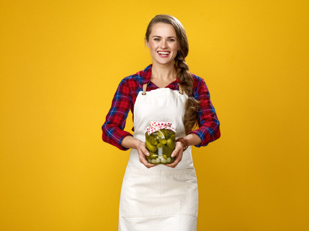 Healthy food to your table. Portrait of smiling young woman farmer wearing apron isolated on yellow showing a jar of pickled cucumbersの写真素材