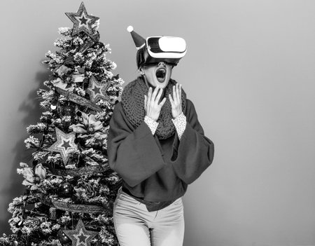 surprised modern woman in colorful clothes near Christmas tree on yellow background wearing virtual...