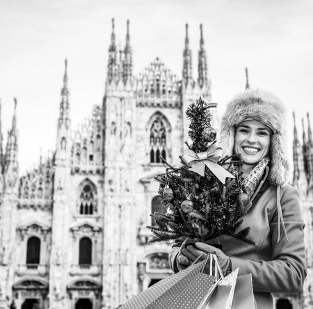 Fun Christmas trip to Milan, Italy. Portrait of happy modern traveller woman in the front of Duomo in Milan, Italy with Christmas tree and shopping bagsの写真素材