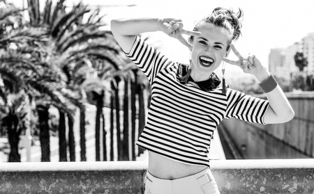 Nosing around, having fun. Full length portrait of happy trendy woman in yellow shorts and stripy shirt against highway in the city showing victory gestureの写真素材